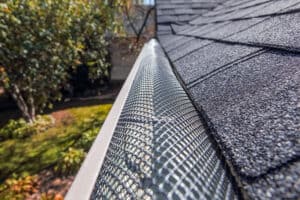 A close-up view of a sloped house roof with black asphalt shingles highlights a metal gutter guard with a mesh design along the edge. In the background, blurred greenery and parts of a tree are visible. For comprehensive care, consider Gutter Services Buchanan County for all your home maintenance needs.
