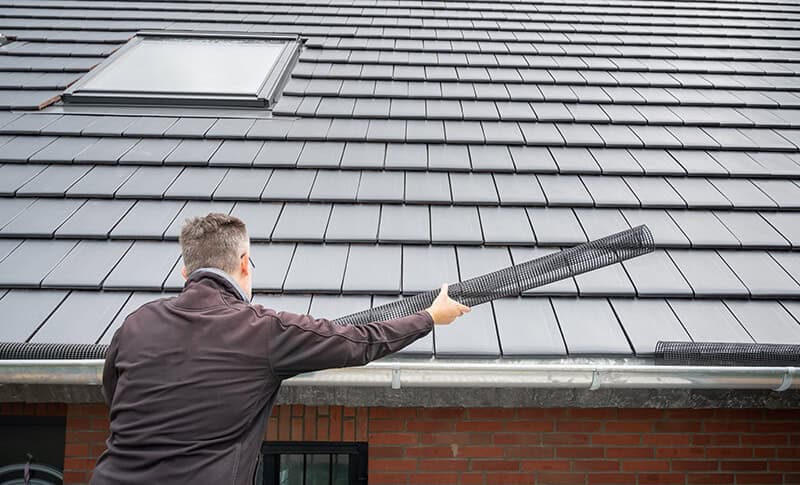 A person in a black jacket is seen from behind while cleaning a home&rsquo;s roof gutter with a long, cylindrical gutter brush. The roof, featuring dark, modern tiles and a skylight, highlights expert roofing in Buchanan County. In the background, part of a brick wall is visible.