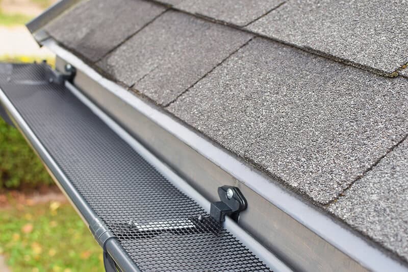 Close-up view of a roof with asphalt shingles and a newly installed black mesh gutter guard. The gutter guard is securely attached along the edge, designed to prevent leaves and debris from clogging the system. This quality installation showcases top-tier Gutter Services in Buchanan County against a leafy backdrop.