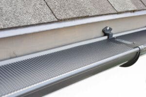 Close-up view of a house gutter system with a protective mesh guard installed by Gutter Services Buchanan County. The mesh prevents debris from clogging the gutter, while the shingle roof and part of the house's fascia are visible above.