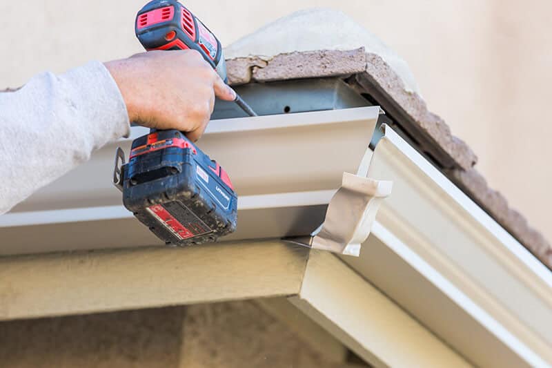 A person is using a red and black power drill to attach a metal gutter to the edge of a roof with brown shingles. The white or light-colored gutter has a piece of tape for alignment. For seamless work like this, trust Gutter Services Buchanan County for all your outdoor home projects.