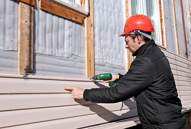 A person wearing a red hard hat and a black jacket uses a power drill to secure siding Buchanan County onto the exterior wall of a building under construction. The wall's insulation and studs are visible, and the worker appears focused on the task.