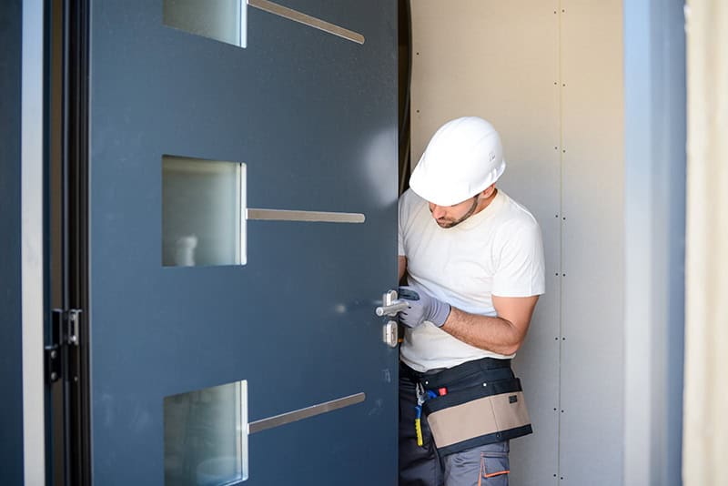 A worker wearing a white hard hat and gloves is installing a modern, dark-colored door with small rectangular windows. He is focused on adjusting the lock, with a tool belt around his waist containing various tools. He stands in a doorway of a building in Buchanan County, where expert door installation services are provided.