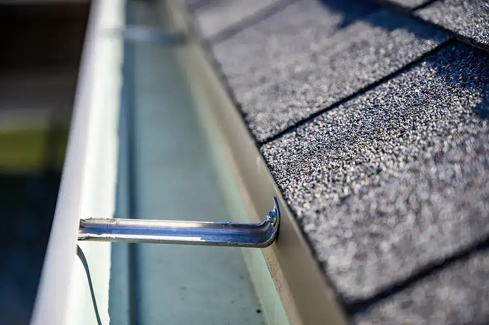 Close-up of a clean, white rain gutter attached to the edge of a roof with grey asphalt shingles. The view highlights a metal bracket securing the gutter to the roof. The texture of the shingles and the smooth gutter are prominent in the sunlight, showcasing expert Gutter Services Buchanan County.