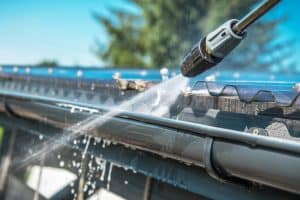A close-up of a pressure washer cleaning the gutter of a house showcases water spraying from the nozzle at high pressure, removing dirt and debris from the metal surface. In the background, a bright blue sky and some blurred greenery can be seen, demonstrating top-notch Gutter Services in Buchanan County.