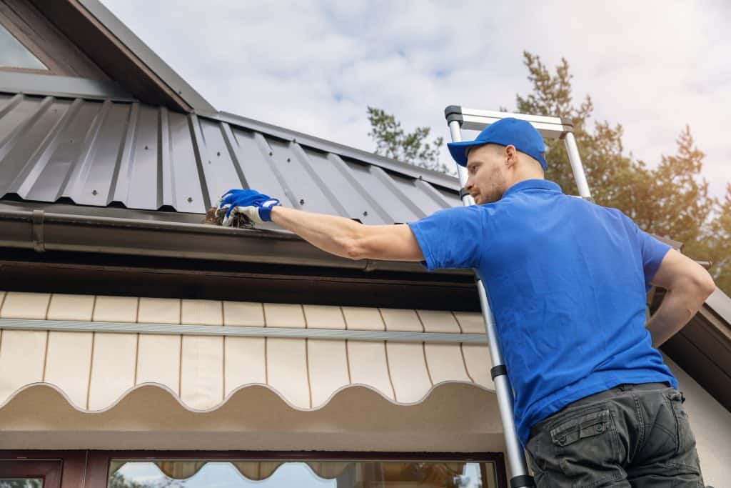 A person wearing a blue shirt, cap, and gloves is standing on a ladder, cleaning out the gutters of a house with a metal roof. The individual is facing away from the camera, holding a clump of debris removed from the gutter. Trees and sky are visible in the background—Gutter Services Buchanan County ensuring clean and efficient drainage.