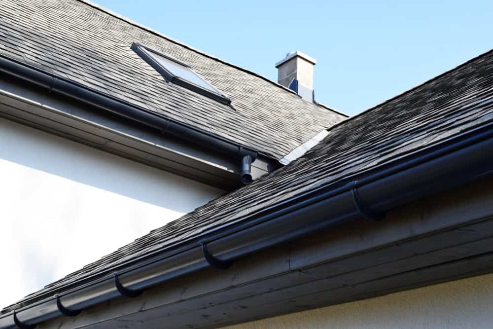 A close-up view of a house roof with gray shingles, featuring a skylight window and a light gray chimney. The roof edge has black rain gutters installed, showcasing professional Gutter Services Buchanan County. The sky in the background is clear and blue.