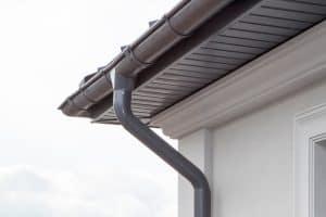 A close-up view of a house’s roof edge in Buchanan County shows a gray rain gutter system attached beneath the eaves. The gutter extends downwards to a matching gray downspout. The light-colored wall contrasts with the cloudy sky in the background, highlighting expert roofing and window installation work.