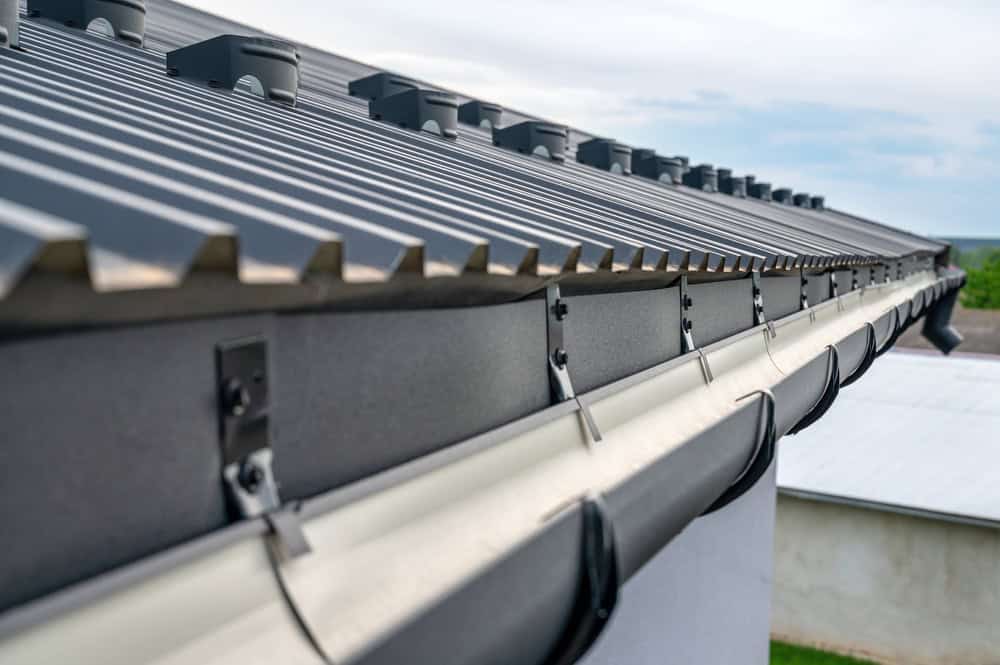 Close-up view of a metal roof with snow guards installed and a black gutter system on the edge. The roof features corrugated steel panels, part of expert Roofing Buchanan County services, and the gutter hangs off the side, ready to collect rainwater. A blurred background shows part of the building and sky.