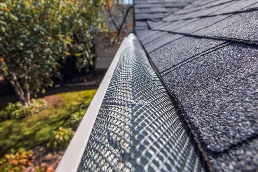 Close-up view of a roof with a shingle covering and a gutter system protected by a metal mesh guard. The background features a landscaped yard with green grass, trees, and some plants. The image highlights the gutter guards' detail and the roof's texture.