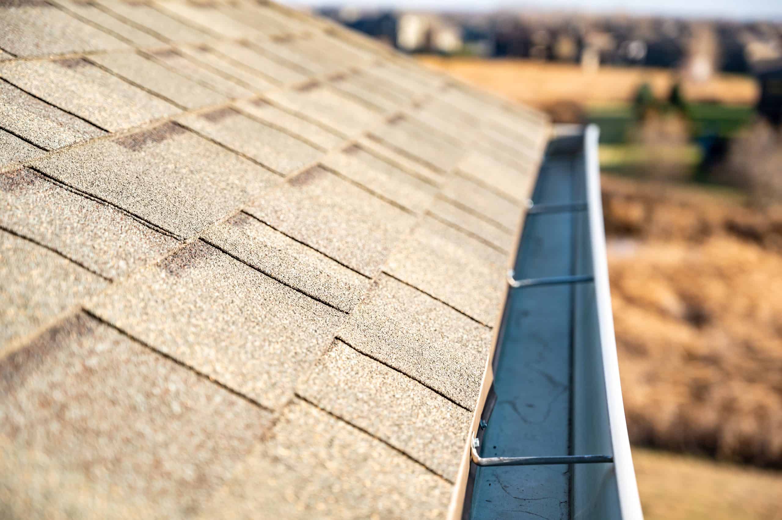 Close-up of an angled rooftop with asphalt shingles and a metal gutter system running along the edge, showcasing expert Gutter Services in Buchanan County. The background is slightly blurred, revealing fields and distant buildings under soft sunlight.