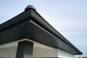 A modern house roof with dark metal soffit and fascia is shown, exemplifying the clean lines of contemporary architecture. The Roofing in Buchanan County highlights a sleek design against a clear sky with light clouds. The sloped roof features a small vent at its peak, complementing the overall elegance.