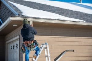 A person wearing a black hoodie and jeans stands on a stepladder fixing a downspout on the side of a house with beige siding in Buchanan County. The roof above is partially covered in snow, and there is a variety of tools attached to the person's tool belt. For all your gutter services needs, trust Buchanan County's experts.