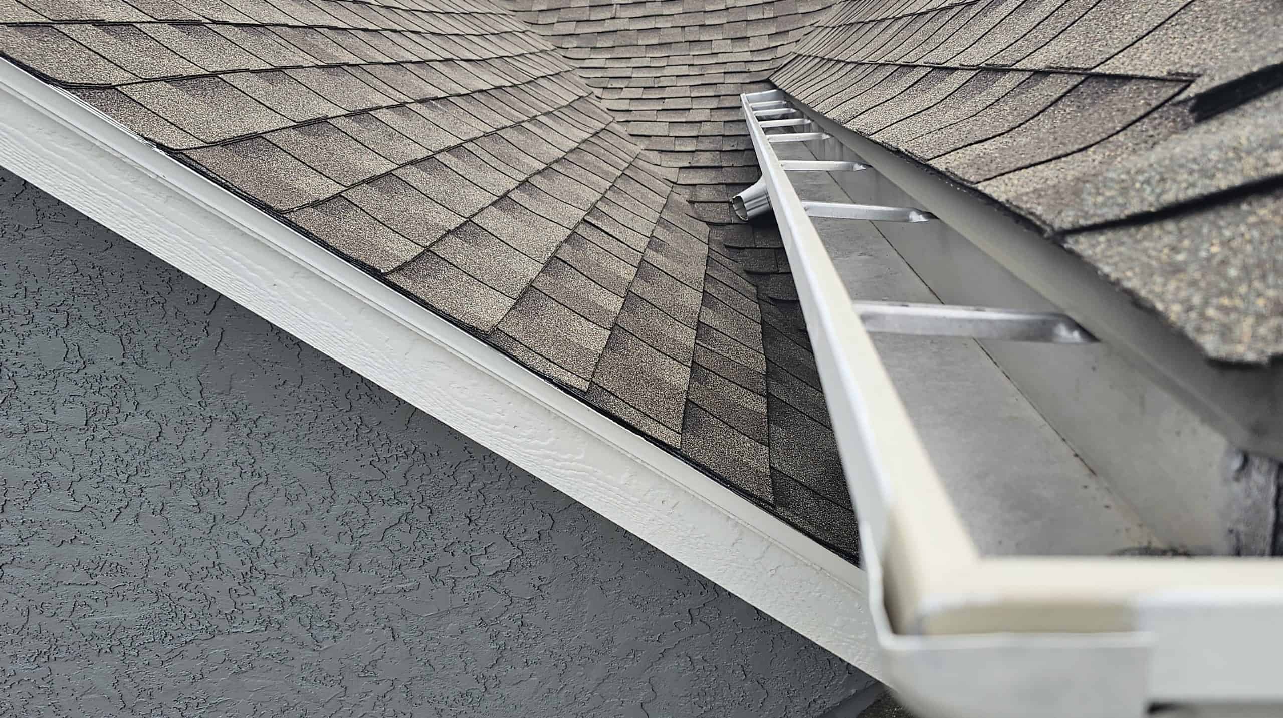 A close-up of a house's rooftop showing two intersecting roof sections with brown shingles. There is a white rain gutter along the edge, exemplifying Gutter Services Buchanan County. The walls are textured with a grey finish, and a silver ladder leans against the gutter.