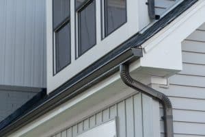 A close-up view of a house's exterior featuring a portion of the roofline, white siding, and a black gutter expertly installed by Gutter Services Buchanan County. The second-story windows are visible above, with black trim and white frames, adding contrast to the multiple layers of siding and wall textures.