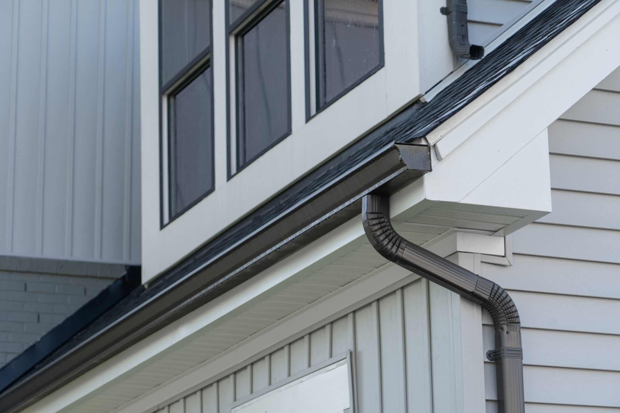 A close-up view of a house's exterior featuring a portion of the roofline, white siding, and a black gutter expertly installed by Gutter Services Buchanan County. The second-story windows are visible above, with black trim and white frames, adding contrast to the multiple layers of siding and wall textures.