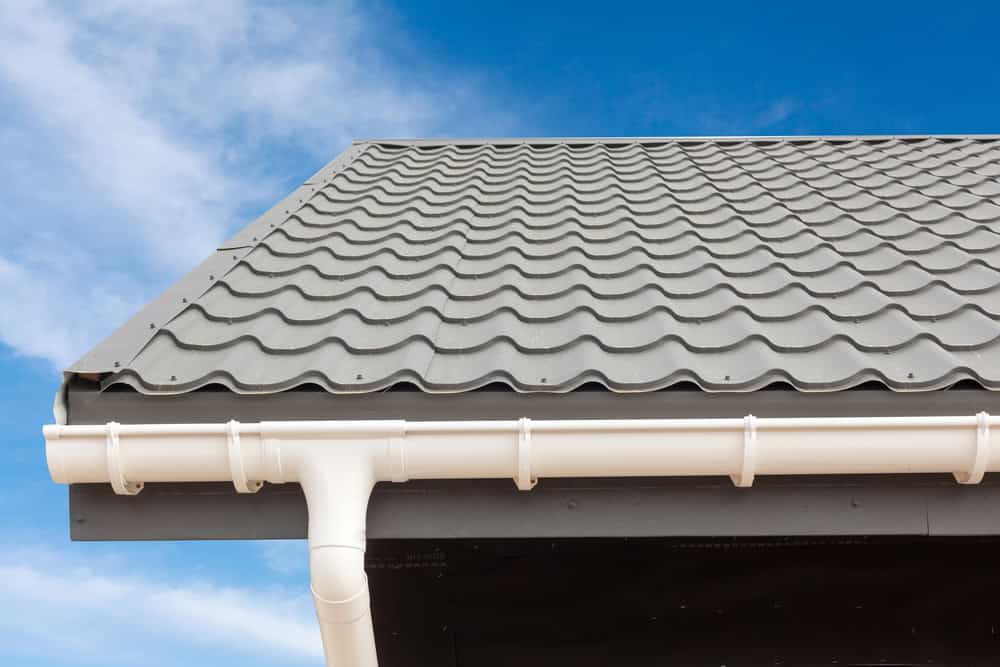 Close-up view of a house roof with grey metal roofing tiles and white gutters against a blue sky. The roof features a wavy tile pattern, and a downspout is seen in the gutter system. For comprehensive home improvements, including gutter services in Buchanan County.
