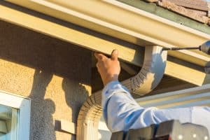 A person on a ladder is installing a beige downspout on the corner of a roof. One hand holds the downspout in place, while the other uses a power drill to secure it. The person, donning a light-colored long-sleeve shirt, is completing this task as part of comprehensive Gutter Services Buchanan County.