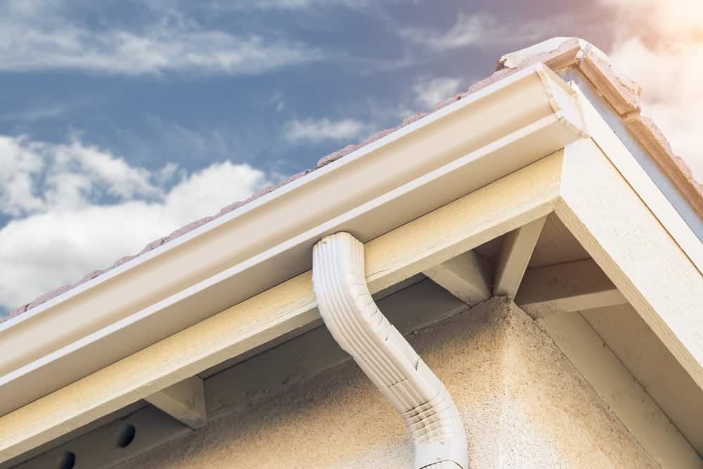 A close-up view of a house's roof eaves, showcasing the gutter and downspout directing rainwater away from the well-maintained roof. The sky in the background is clear with some scattered clouds. Roofing Buchanan County ensures light-colored paint and roofing materials remain pristine.
