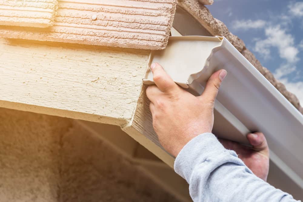 Two hands are installing a white rain gutter on the edge of a house roof with textured, brown shingles. In the background, the sky is partially visible as they work. This meticulous attention to detail mirrors similar quality services for window installation in Buchanan County.