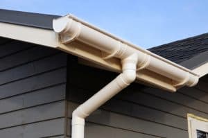 A close-up view of a house corner in Buchanan County features a beige rain gutter and downspout system attached to a roof with dark shingles. The siding, also in Buchanan County, is dark gray, contrasting nicely with the clear blue sky in the background.
