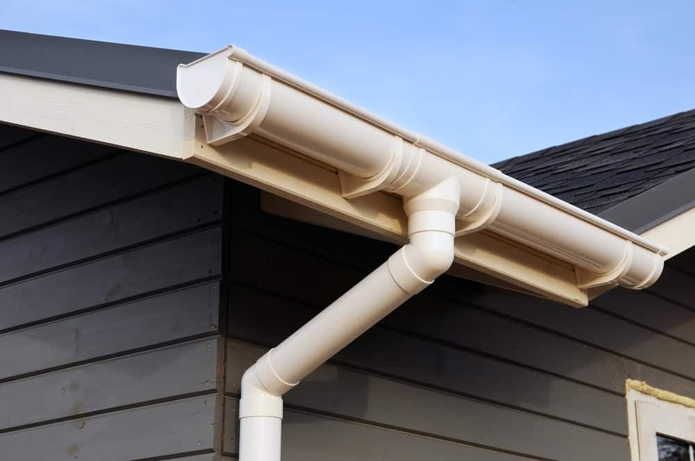 A close-up view of a house corner in Buchanan County features a beige rain gutter and downspout system attached to a roof with dark shingles. The siding, also in Buchanan County, is dark gray, contrasting nicely with the clear blue sky in the background.