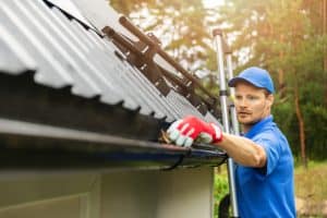 A man wearing a blue shirt, cap, and red gloves stands on a ladder next to a house, cleaning out the gutter with his hand. The roof is made of corrugated metal and the background features a forested area. The man is focused on his task.