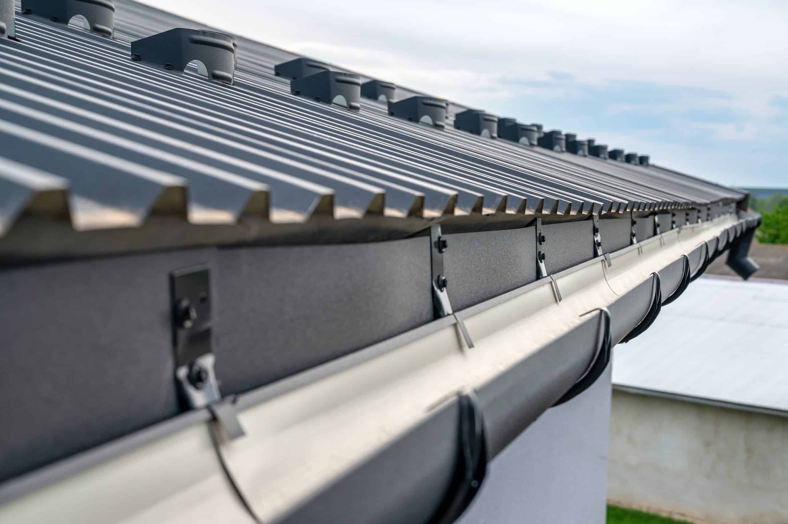 Close-up view of a modern metal roof with a gutter system attached to the edge of a building, showing the corrugated metal panels and downspout brackets under a cloudy sky.