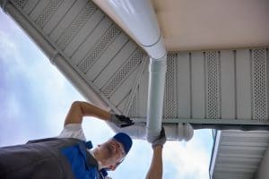 A worker in a blue hat and uniform is fixing a white gutter under the roof of a house. The photo is taken from below, showing the underside of the roof and the man reaching up to adjust the gutter. The sky is visible in the background.