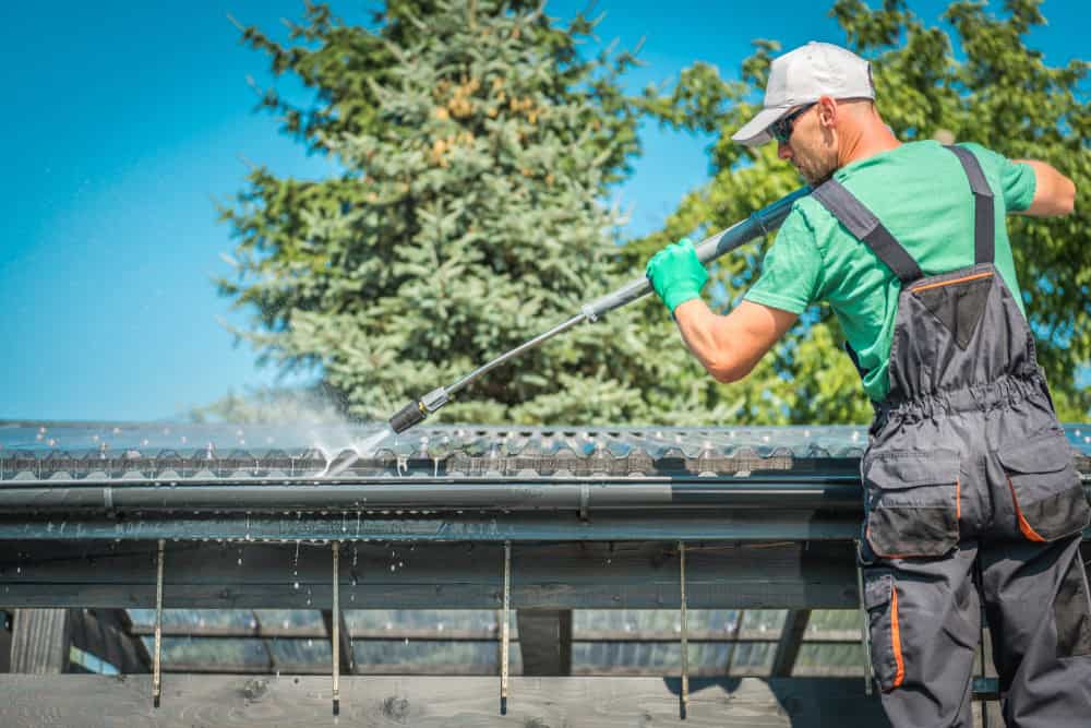 A worker in a green shirt, gray overalls, and green gloves power washes the roof of a building. He stands on a ladder, using a high-pressure hose to clean the roof, with a large pine tree and a clear blue sky in the background.