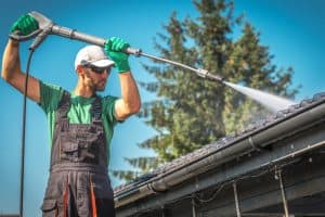 A man wearing protective gloves, sunglasses, a green shirt, and overalls uses a high-pressure water sprayer to clean the roof of a building. The background includes a clear blue sky and a tall tree.