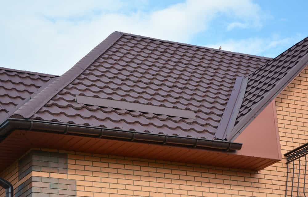 A close-up of a house roof with brown metal roofing tiles and brick walls. The roof has a gable design, and the metal tiles are uniformly arranged. The house exterior includes various shades of beige and brown bricks, creating a patterned look.