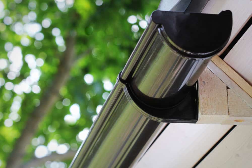 Close-up of a modern black gutter attached to the edge of a roof. The gutter is mounted on a wooden structure, and the background features blurred green leaves and tree branches, indicating an outdoor setting.