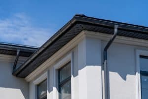 Close-up view of a modern house corner with white walls and large windows, showcasing black roof gutters and downspouts against a clear blue sky. The structure is well-defined with sharp lines and shadows, highlighting the building's contemporary design.