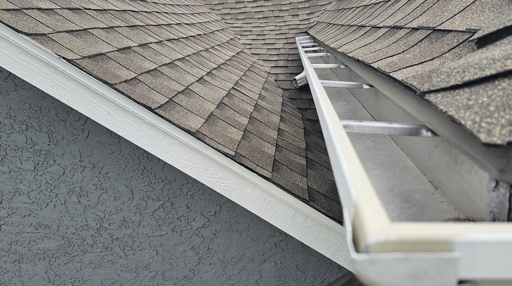 A close-up view of a house roof with asphalt shingles. A ladder leans against the roof, and a white rain gutter runs along the roof edge. The gutter is clean, and the shingles appear to be in good condition.