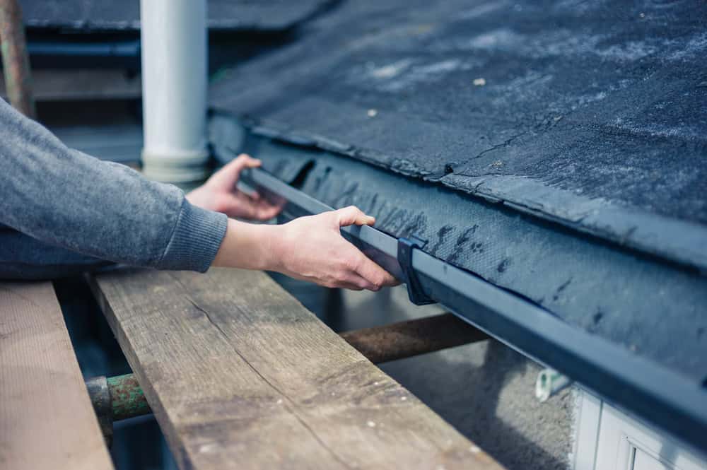 Hands of a person in a grey sweater fixing a black plastic gutter on the roof of a building. The person is standing on a wooden scaffold platform. The roof appears to have weathered, exposed portions and the setting suggests an outdoor maintenance activity.