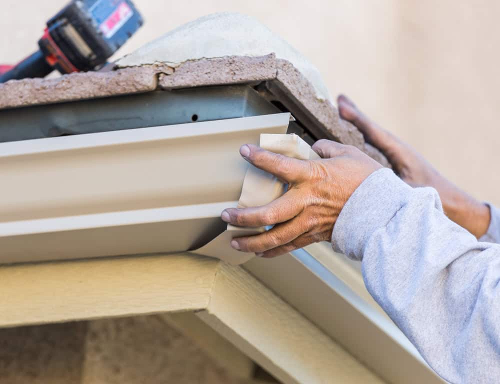 Close-up of a person’s hands wearing a long-sleeved shirt, installing or adjusting a piece of white metal guttering at the edge of a roof. Tools rest on the roof above the gutter. The setting appears to be a home exterior.