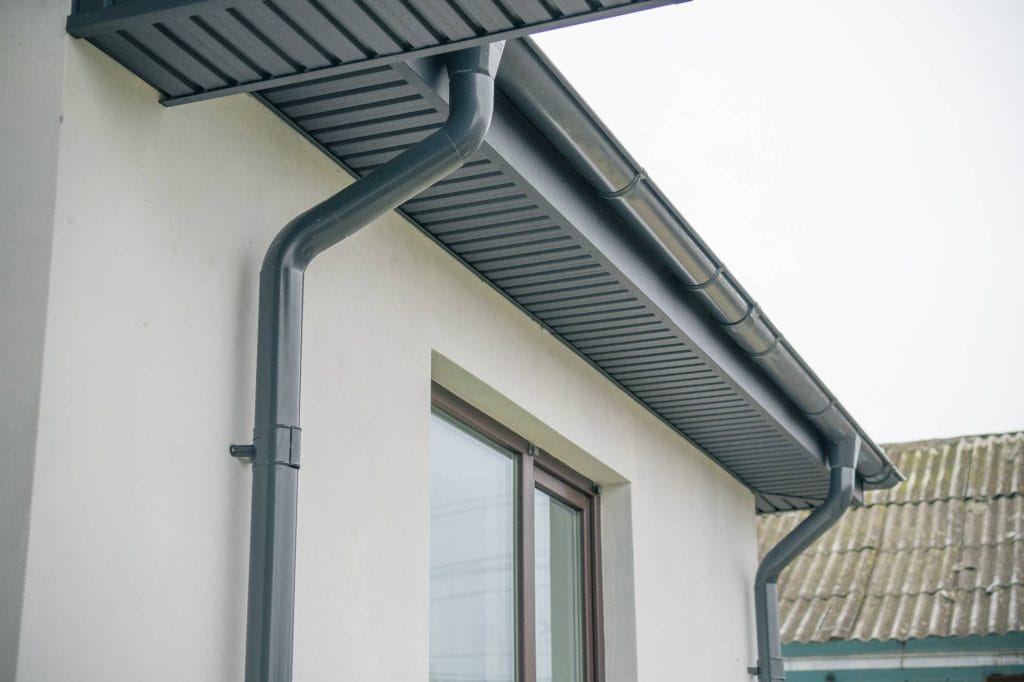 Close-up of a gray rain gutter and downspout attached to the roof of a light-colored house, with a window and part of a corrugated roof visible in the background.