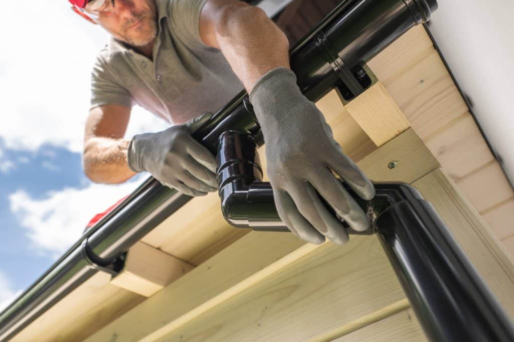 A man wearing gloves installs a black rain gutter on the edge of a wooden roof, securing the corner section under a blue sky.
