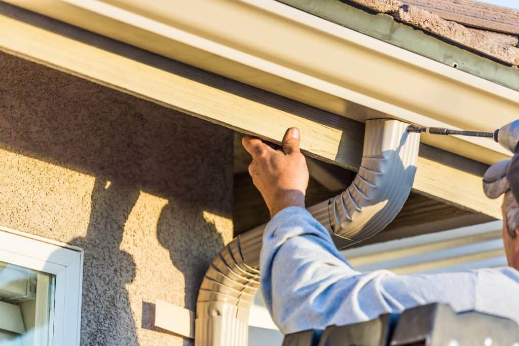 A person on a ladder installs or repairs a house gutter system, securing a downspout near the roofline on a sunny day.
