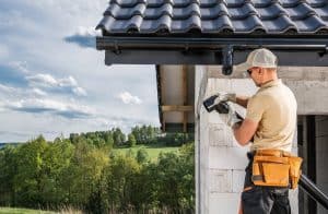 A construction worker wearing a tool belt, gloves, and a cap uses a power drill on the exterior wall of a house under construction, with a tiled roof and a scenic, green landscape in the background.