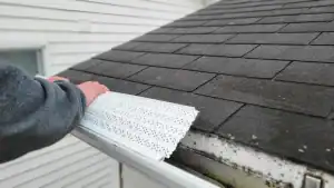 A person’s hand installs a perforated gutter guard on a house roof with dark shingles and white siding.