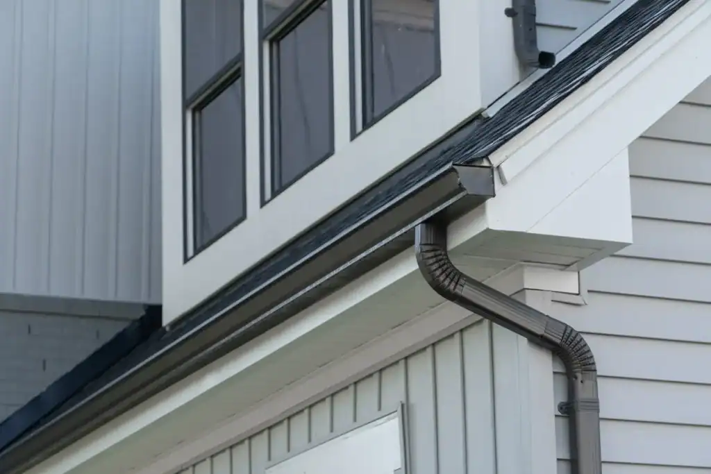 Close-up of a house exterior showing grey siding, white trim, black rain gutter and downspout, and several large windows on the upper floor reflecting the sky.