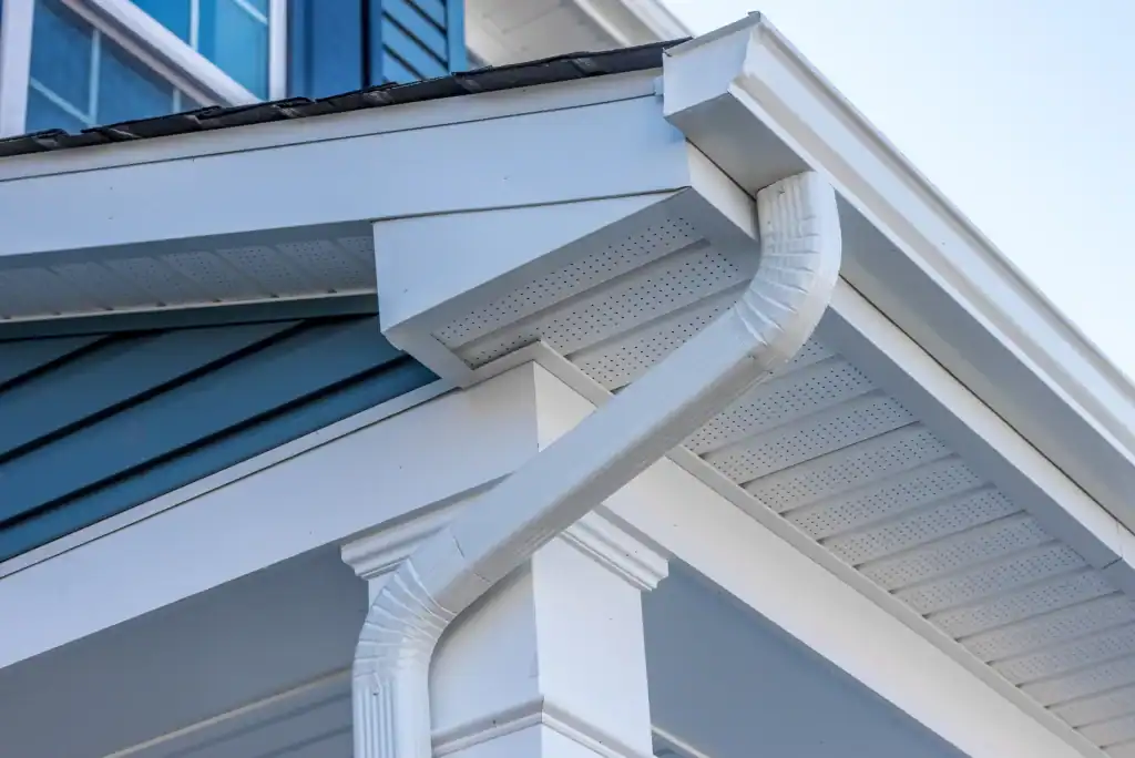 Close-up of a house corner showing a white roof gutter and downspout system attached to the roof edge, with blue siding and white trim visible in the background.