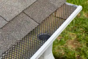 Close-up of a house roof with brown shingles, a white gutter, and a black mesh guard covering the gutter to prevent debris from entering; green grass is visible in the background.