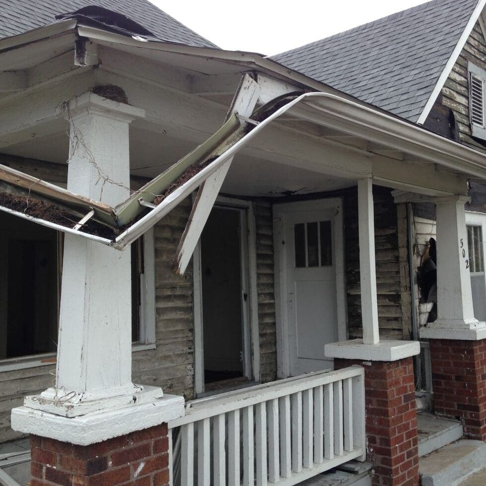 A house with severe damage to the front porch, including a collapsed roof and broken columns, showing exposed wood and missing panels. The open door adds to its unsafe appearance&mdash;ideal for those in need of urgent home services.